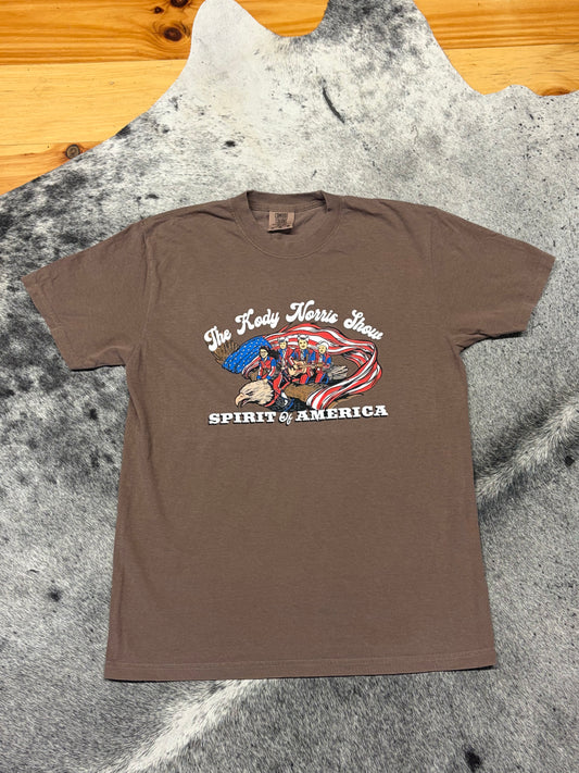 A brown graphic t-shirt laid flat on a gray and white cowhide rug, with a wooden floor visible in the top left corner. The shirt features a patriotic graphic with the text 'The Lady Horse Show' and 'SPIRIT OF AMERICA' surrounding an eagle and American flag design.