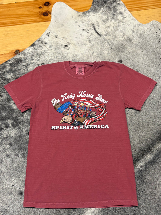 A maroon short-sleeve graphic t-shirt laid flat on a gray and white cowhide rug, with a wooden floor visible in the top left corner. The shirt features a graphic of a Native American holding a flag with the text 'The Redskin Morris Show' and 'SPIRIT OF AMERICA'.