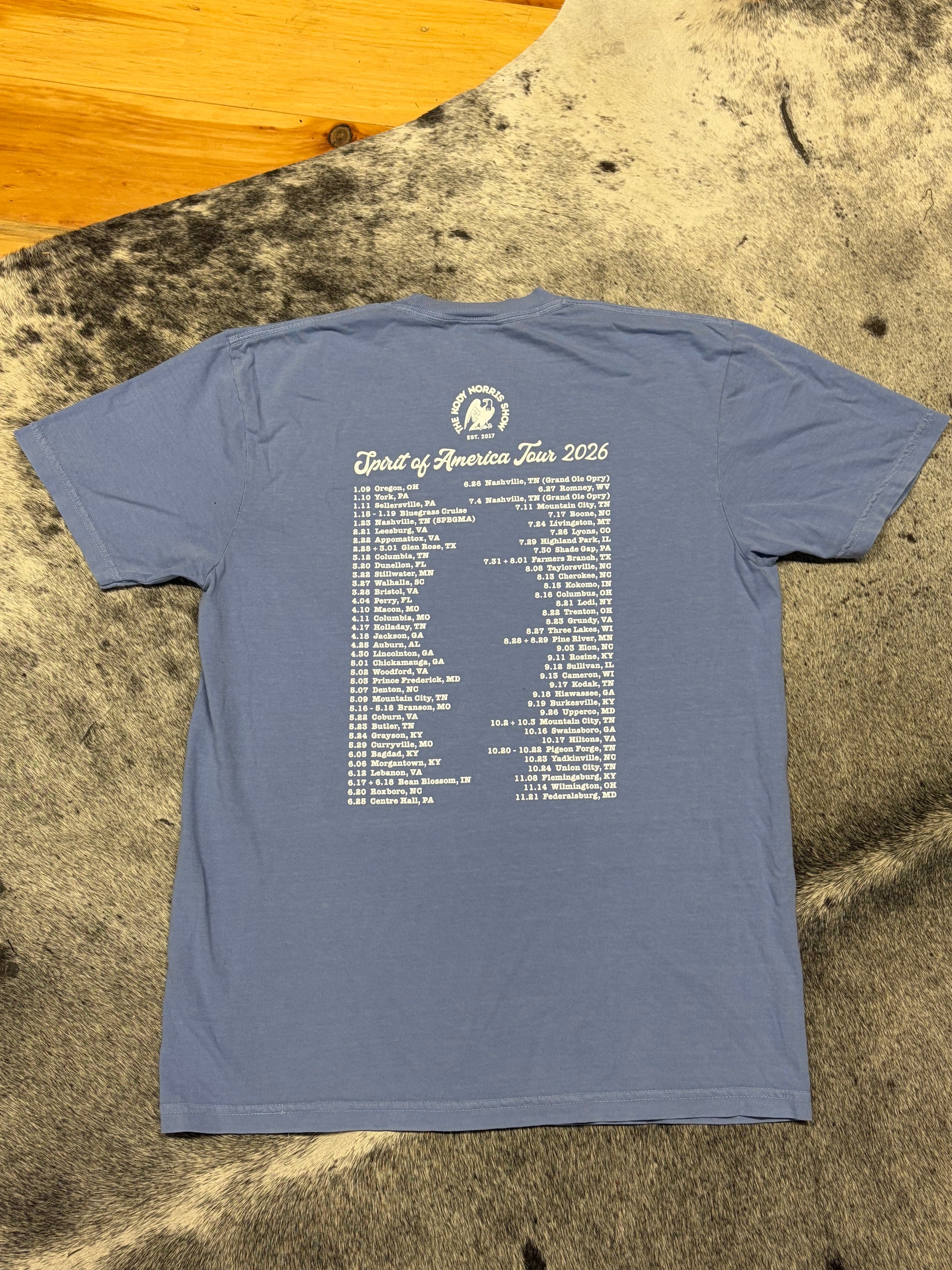 A slate blue t-shirt laid flat on a textured cowhide rug, showing the back of the shirt with a white printed list of 'South of America Tour 2026' dates and a circular logo.