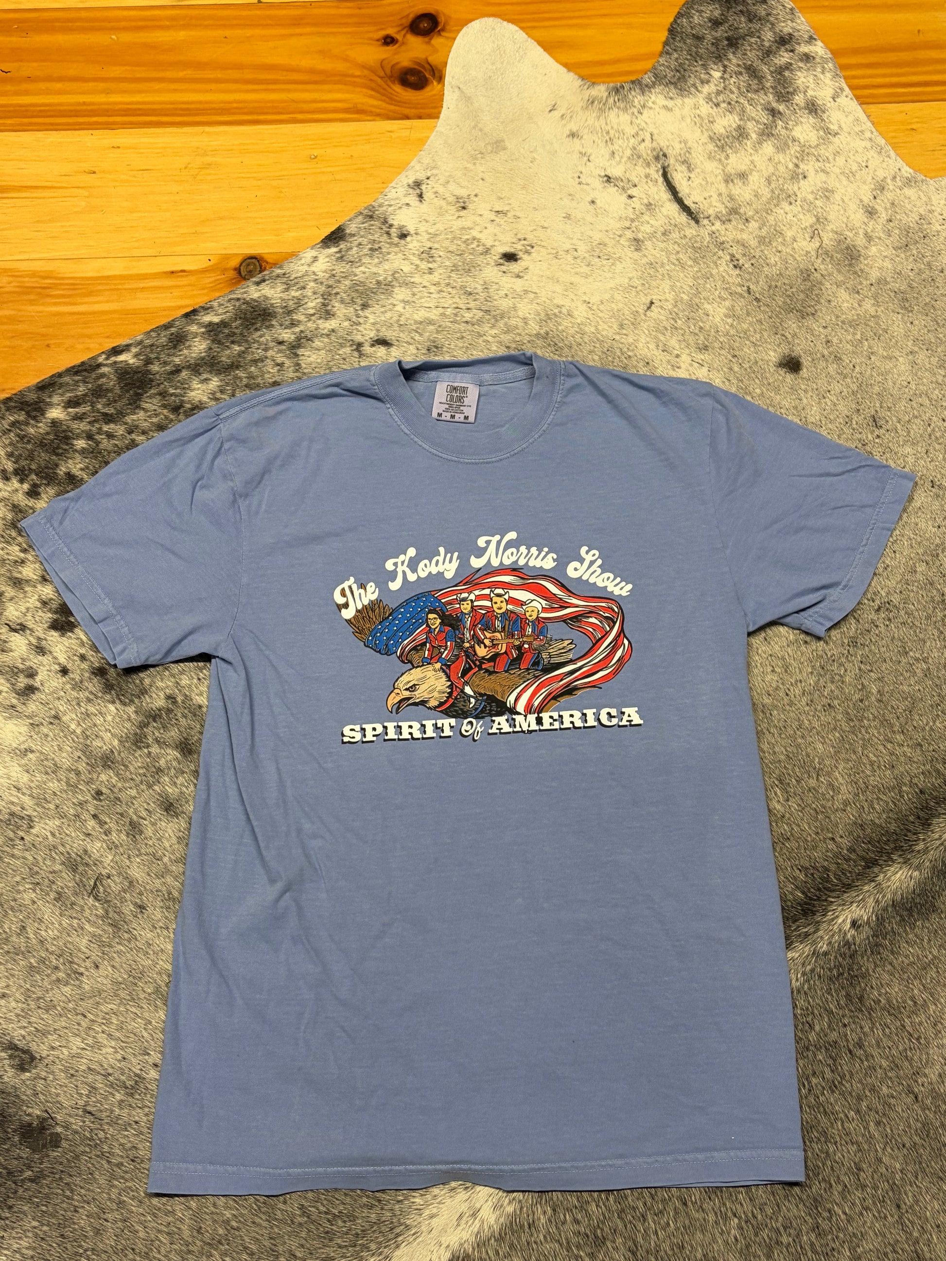 A light blue graphic t-shirt laid flat on a black and white cowhide rug over a wooden floor. The graphic features a Native American head with an eagle and a waving American flag, with the text 'The Red Horse Show' and 'SPIRIT OF AMERICA'.